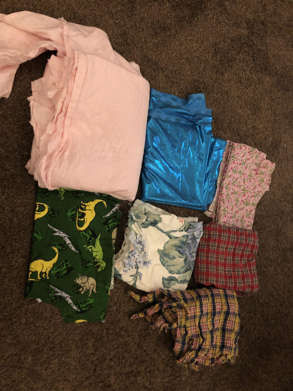 Color photo of 7 folded piles of fabric on a brown carpet- 1 pink flannel, 1 blue ycra, 1 pink flower-printed, 1 red plaid, 1 yellow and blue plaid, 1 white floral, and 1 green with yellow dinosaurs